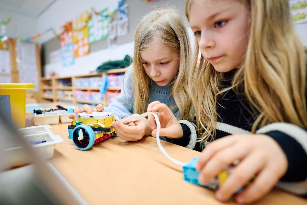 Zwei Kinder sitzen an einem Klassentisch und bauen Roboterbausätze zusammen, umgeben von bunten Materialien.
