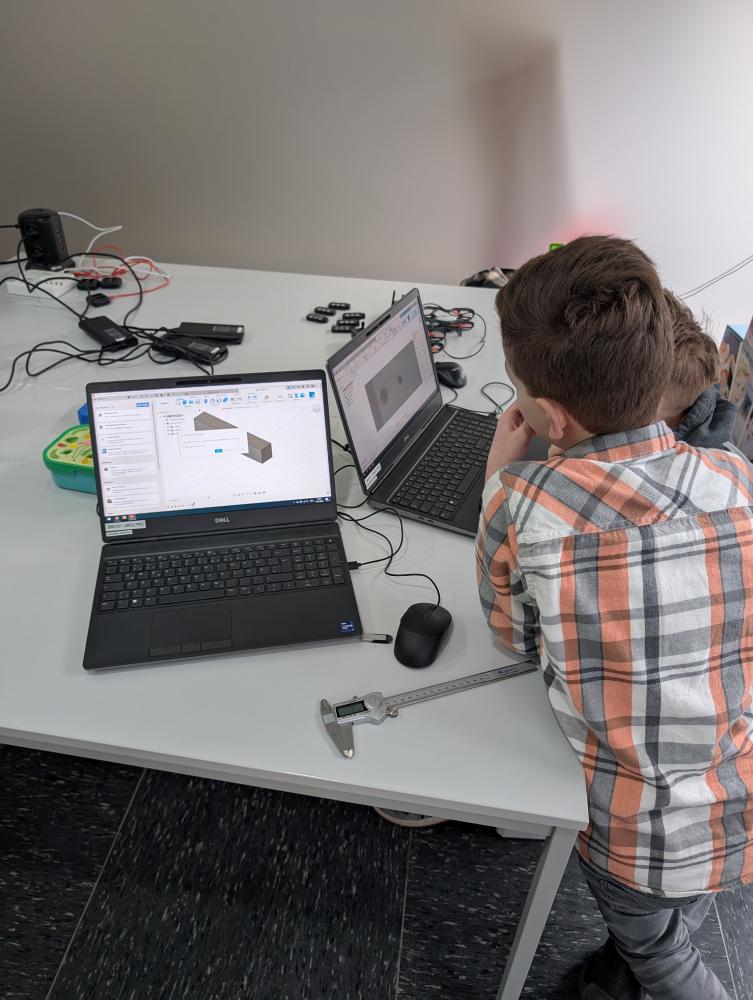 Ein Kind arbeitet an einem Laptop, der auf einem Tisch steht. Ein weiterer Laptop steht in der Nähe. Auf dem Tisch liegt außerdem ein Messschieber und verstreute elektronische Bauteilen.