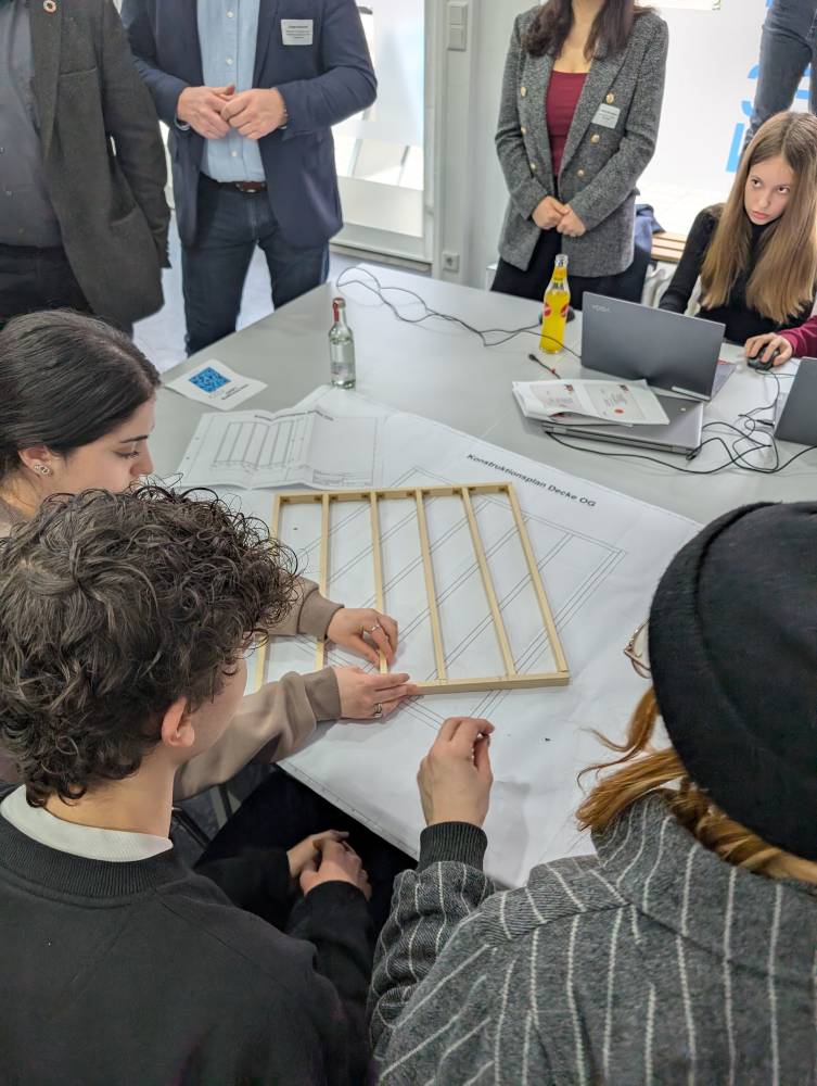 Eine Gruppe von Menschen sitzt um einen Tisch herum und arbeitet gemeinsam an einem Projekt mit Holzstäben und großen Blättern Papier. Einige Personen beobachten die Aktivität. Auf dem Tisch stehen auch Laptops und Flaschen.