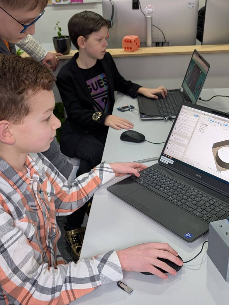 Zwei Kinder sitzen an einem Tisch und arbeiten jeweils an einem Laptop. Ein Kind schaut den beiden Kindern über die Schulter auf den Bildschirm des Laptops.
