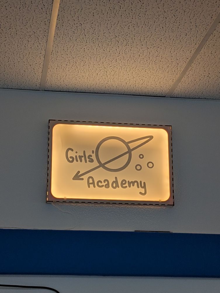 Das Bild zeigt ein beleuchtetes Schild an einer Wand mit der Aufschrift „Girls‘ Academy“. Neben dem Text ist ein Planet mit Ringen und einem nach links zeigenden Pfeil gezeichnet.