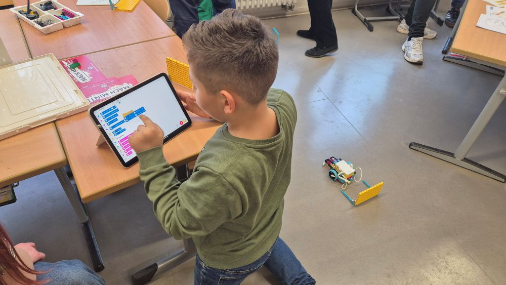 Ein Kind im grünen Pullover sitzt an einem Tisch und programmiert mit einem Tablet einen kleinen Roboter auf Rädern, der auf dem Boden steht.