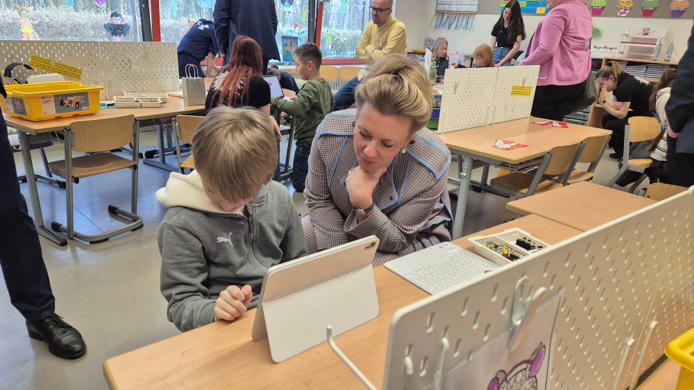 Eine Frau sitzt mit einem Kind an einem Tisch und schaut auf ein Tablet. Im Hintergrund sind andere Personen zu sehen, die ebenfalls an Tischen beschäftigt sind.