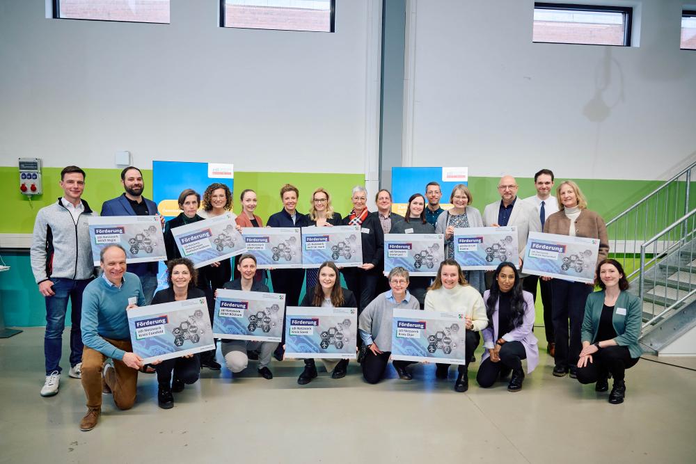Eine Gruppe von Menschen steht in einem Gebäude, hält in zwei Reihen große Schilder in den Händen und lächelt in die Kamera.
