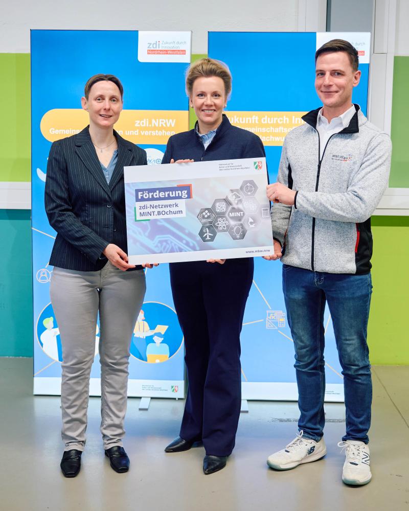 Drei Personen stehen vor zwei blauen Aufstellern und halten ein Plakat zur Förderung des zdi-Netzwerks MINT.BOchum in den Händen.