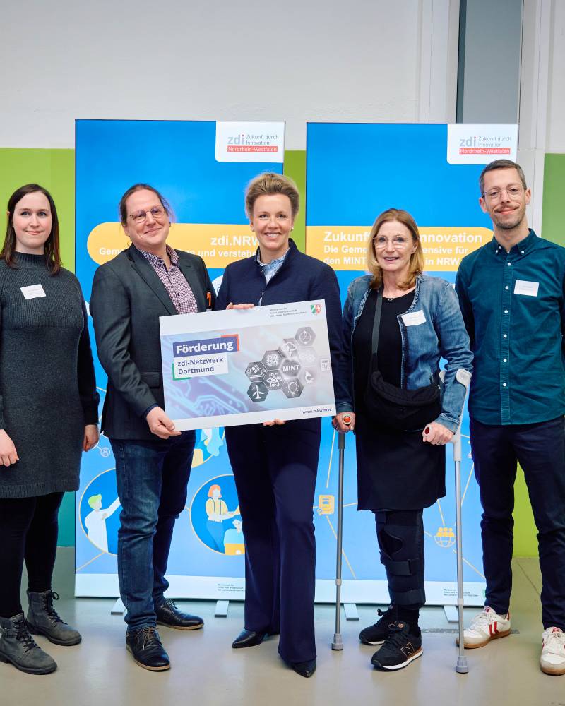 Fünf Personen stehen zusammen und halten ein großes Schild mit der Aufschrift "Förderung zdi-Netzwerk Dortmund". Im Hintergrund sind zwei blaue Banner mit Logos zu sehen.