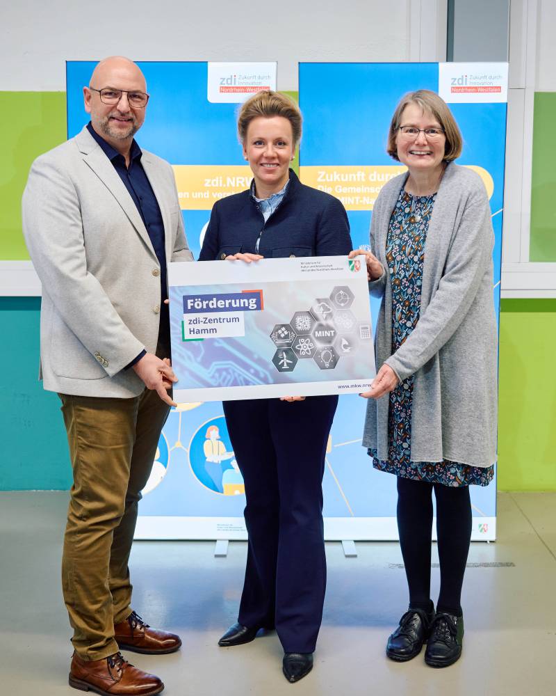 Drei Personen stehen vor zwei blauen Aufstellern und halten ein Schild mit der Aufschrift „Förderung zdi-Zentrum Hamm“ in den Händen.