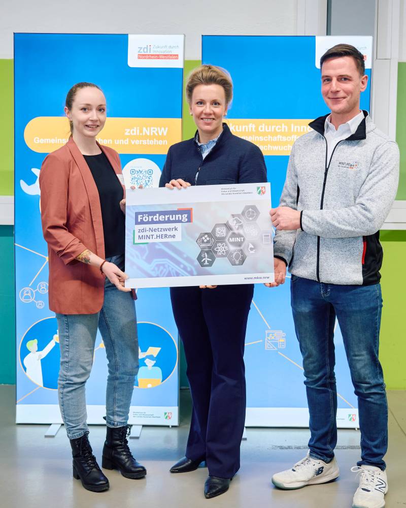 Drei Personen stehen vor zwei blauen Aufstellern und halten ein Schild mit der Aufschrift „Förderung zdi-Netzwerk MINT.HERne" in den Händen.