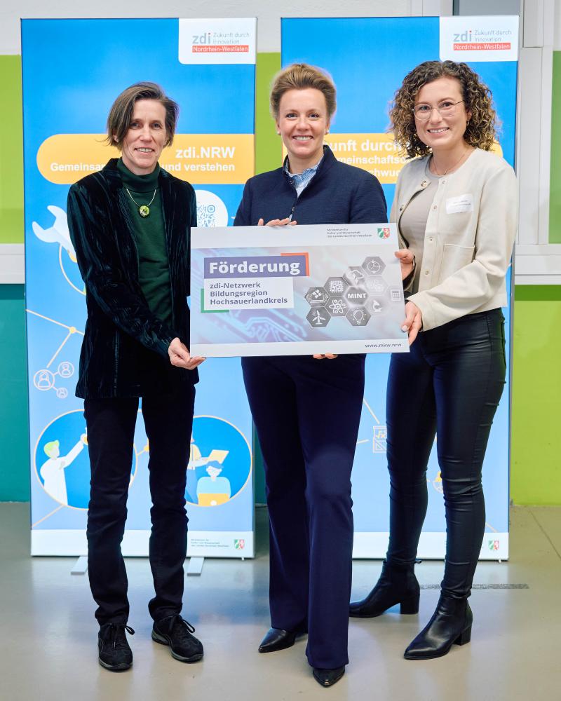 Drei Personen stehen vor zwei blauen Aufstellern und halten ein Schild mit der Aufschrift „Förderung zdi-Netzwerk Bildungsregion Hochsauerlandkreis“ in den Händen.