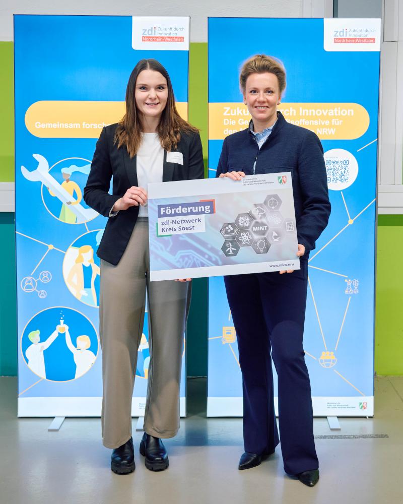 Zwei Personen stehen auf einem hellen Boden und halten ein Schild mit der Aufschrift "Förderung zdi-Netzwerk Kreis Soest" vor zwei blauen Bannern. Eine Person trägt eine schwarze Jacke und eine helle Hose, während die andere Person einen marineblauen Anzug trägt.