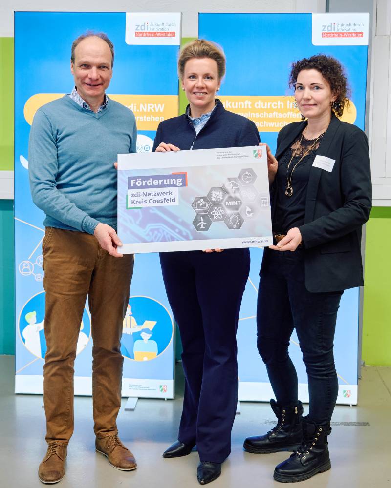 Drei Personen halten ein großes Schild mit der Aufschrift "Förderung zdi-Netzwerk Kreis Coesfeld in der Hand. Hinter ihnen sind blaue Banner mit Logos und Text zu sehen. Sie tragen legere und Business-Casual-Kleidung.