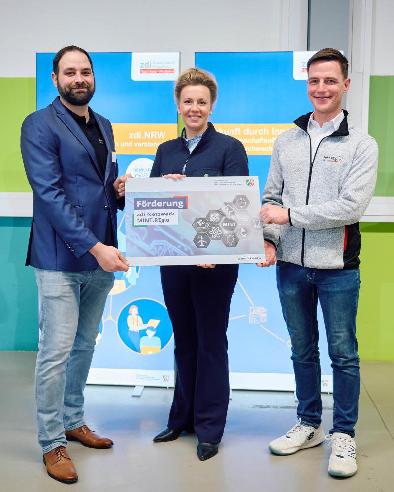 Drei Personen stehen vor zwei blauen Aufstellern und halten ein Schild mit der Aufschrift „Förderung zdi-Netzwerk MINT.REgio“ in den Händen.