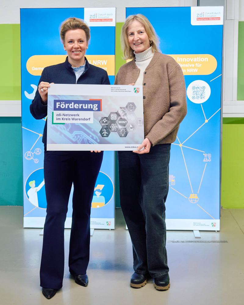 Zwei Personen stehen vor zwei blauen Aufstellern und halten ein Schild mit der Aufschrift „Förderung zdi-Netzwerk im Kreis Warendorf“ in den Händen.