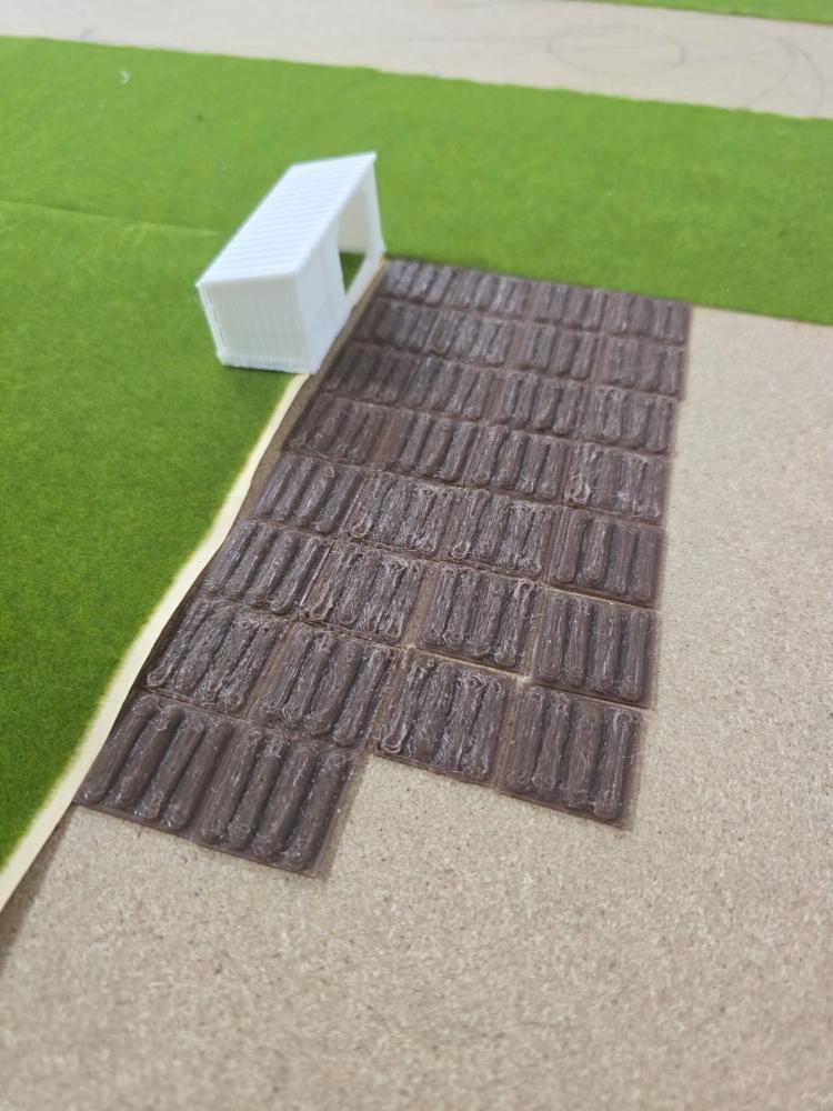 Das Bild zeigt ein maßstabsgetreues Modell eines kleinen weißen Bauwerks auf einer Rasenfläche, zu dem ein braun gefliester Weg führt.