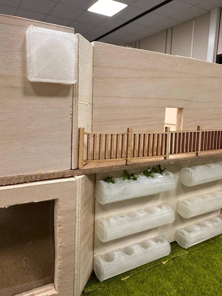 Das Bild zeigt ein hölzernes Architekturmodell mit Kunststoffelementen, darunter ein Zaun und kleine Pflanzen auf einer Seite, alles auf einer grasähnlichen grünen Oberfläche platziert.