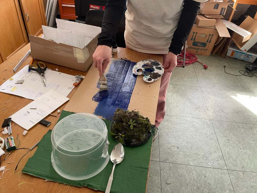 Eine Person bemalt ein Holzbrett blau an einem überladenen Tisch, der mit Künstlerbedarf wie einer Palette, einem Pinsel und Moos bedeckt ist.