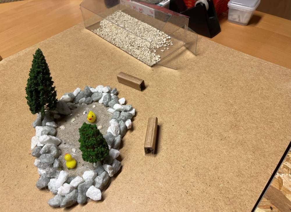 Auf einer Tischplatte befindet sich eine Miniaturszene mit zwei kleinen Bäumen und einem felsigen Teich mit zwei Gummienten. Zum Aufbau gehören außerdem zwei kleine Baumstämme und ein durchsichtiger Behälter mit winzigen Kieselsteinen.