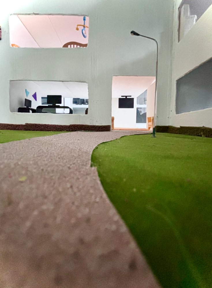 Das Bild zeigt ein Miniaturmodell eines modernen Gebäudes mit großen Fenstern. Es ist von einer grünen Wiese und einem Weg umgeben und verfügt über eine Straßenlaterne. Im Inneren befindet sich ein Arbeitsbereich mit Stühlen und Schreibtischen.