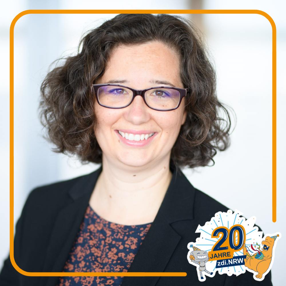 Eine Frau mit braunen Locken und Brille lächelt in die Kamera und trägt einen Blazer. Unten rechts im Bild prangt ein „20 Jahre zdi.NRW“-Button.