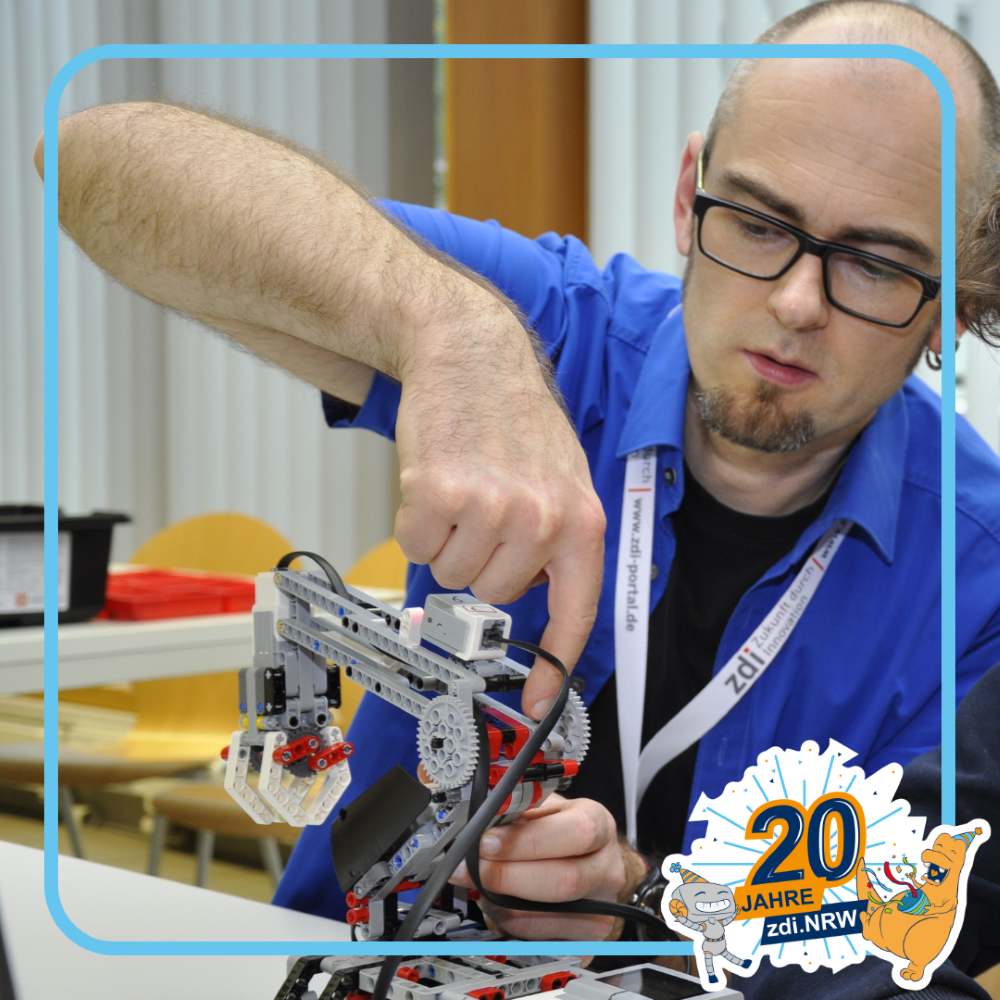 Ein Mann mit Brille und blauem Hemd justiert ein Robotergerät auf einem Tisch. In der unteren rechten Ecke prangt ein „20 Jahre zdi.NRW“-Schild.