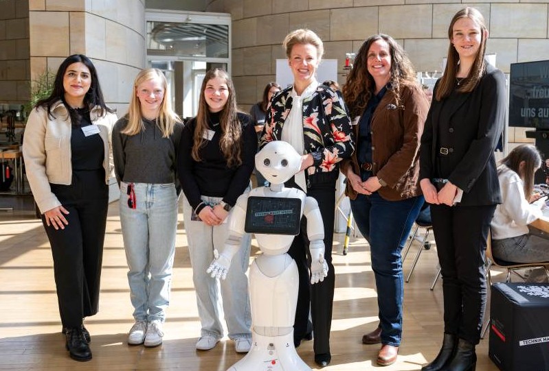 Eine Gruppe von sechs Personen, darunter ein humanoider Roboter, posiert für die Kamera. Die lächelnde Gruppe besteht aus zwei jungen Mädchen und vier erwachsenen Frauen, die sich um den Roboter versammelt haben.