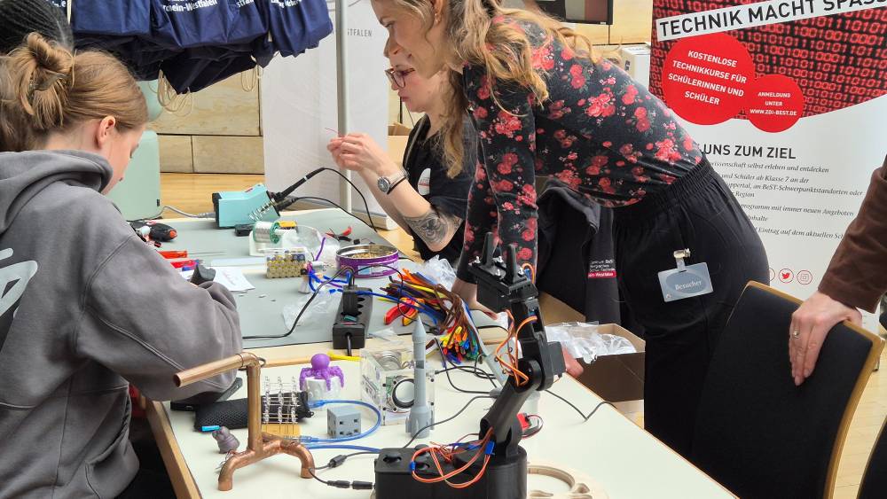 In einer geschäftigen Werkstatt arbeiten Menschen an einem Tisch voller elektronischer Geräte und Roboterkomponenten. Umgeben von Kabeln und Werkzeugen bereiten sie sich auf den Zukunftstag 2025 vor, mit dem Ziel grenzenloser Innovation.