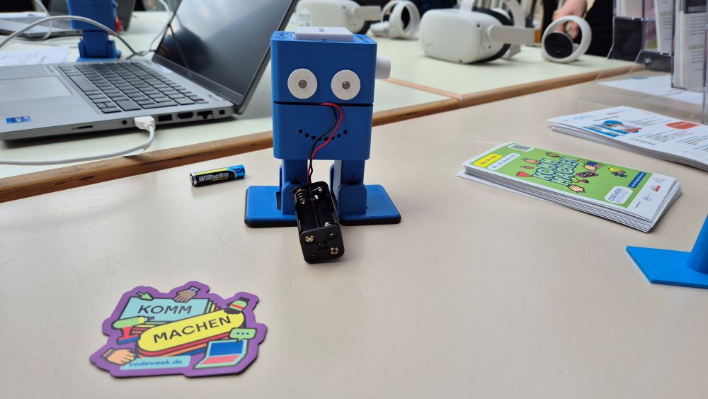 Ein kleiner blauer Roboter mit Wackelaugen sitzt auf einem Tisch neben einem Laptop, einem Akkupack und Broschüren. Auf dem Tisch steht ein Aufkleber mit der Aufschrift „Komm Machen“.