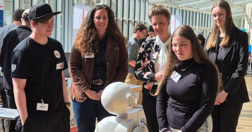 Beim Zukunftstag 2025 beschäftigt sich eine Gruppe von Menschen bei einer Indoor-Veranstaltung mit einem kleinen humanoiden Roboter und erkundet die Zukunft der Technologie.