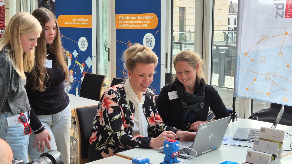 Eine Gruppe von Menschen sitzt mit Laptops um einen Tisch herum. Im Hintergrund hängen leuchtend bunte Banner.