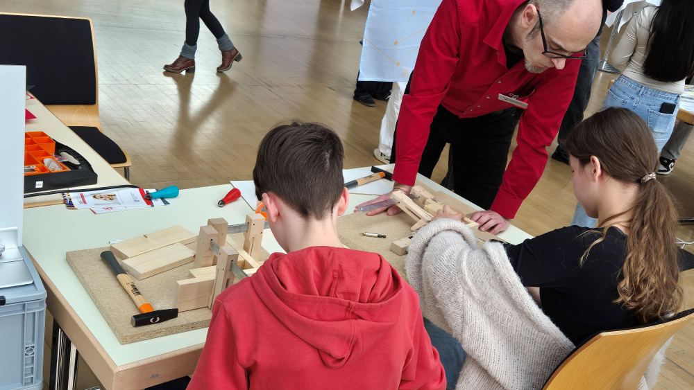Zwei Kinder bauen an einem Tisch Holzkonstruktionen zusammen, angeleitet von einem Erwachsenen im roten Hemd. Im Hintergrund sind weitere Personen zu sehen.