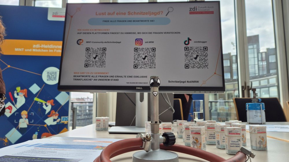 Auf einem Computerbildschirm wird eine digitale Schnitzeljagd mit QR-Codes zum Zukunftstag 2025 gezeigt. Davor steht ein wissenschaftliches Gerät auf einem Tisch, umgeben von kleinen Behältern, wodurch eine faszinierende Mischung aus Technologie und Wissenschaft in einem pädagogischen Umfeld entsteht.