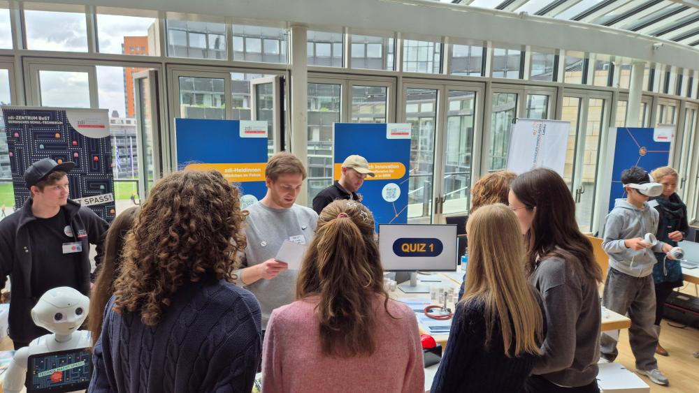 Auf der Tech-Ausstellung „Zukunftstag 2025“ versammeln sich die Menschen um einen mit „QUIZ!“-Plakaten dekorierten Stand. Sie sind fasziniert von Robotern und hochmodernen Geräten, die auf dem Tisch ausgestellt sind. Große Fenster im Hintergrund geben den Blick auf eine weite Stadtlandschaft frei.