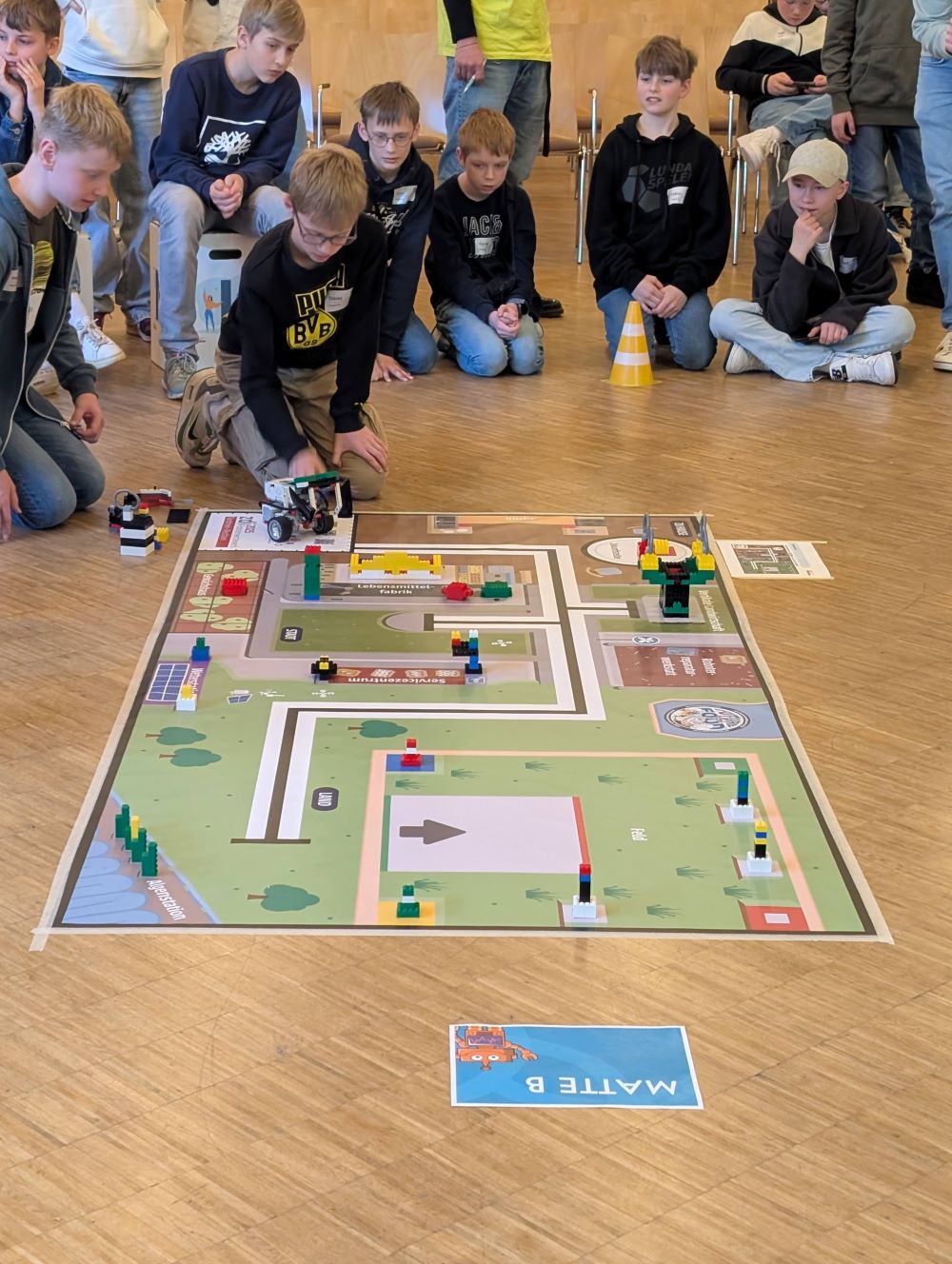 Mehrere Schüler knien auf dem Boden vor einer Spielfeldmatte. Zwei Jungs bereiten einen Legoroboter für einen Lauf beim zdi-Roboterwettbewerb vor.
