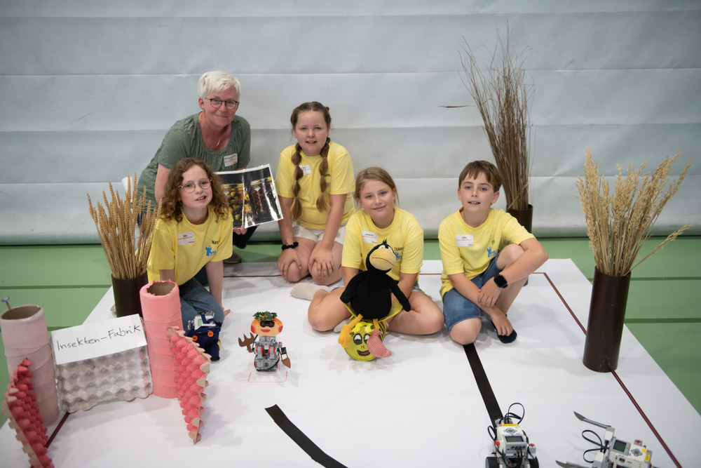 Eine Gruppe von Kindern und ein Erwachsener sitzen in einer Turnhalle auf dem Boden. Sie haben kleine Roboterprojekte vor sich liegen. Bastelmaterial und Pflanzen sind um sie herum verstreut.