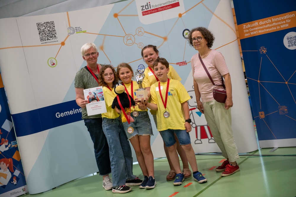 Fünf Personen stehen in einem Raum und posieren für ein Gruppenfoto bei einer Bildungsveranstaltung. Drei Kinder tragen Medaillen und halten Urkunden, während eine Person einen Pokal in der Hand hält.