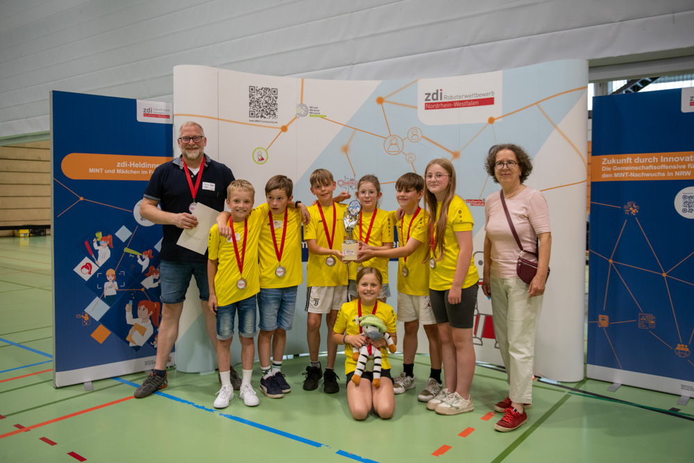 Eine Gruppe von Kindern in gelben Hemden und mit Medaillen posiert mit zwei Erwachsenen und einem Pokal bei einer Hallenveranstaltung. Im Hintergrund sind Transparente zu sehen.