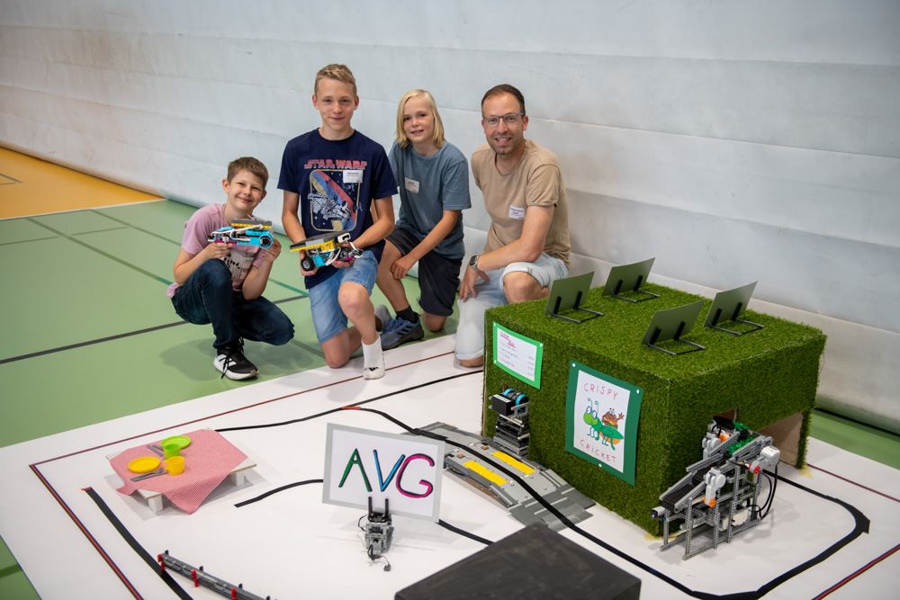 Drei Jungen und ein Erwachsener knien auf dem Boden einer Turnhalle neben einem Modellroboterprojekt. Das Projekt besteht aus beschrifteten Schildern, Spielzeugnahrung, einer grünen Kunstrasenstruktur und Laptops obenauf.