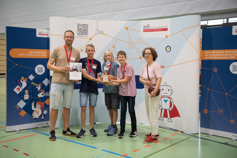 Vier Kinder und zwei Erwachsene stehen drinnen mit einem Pokal und einer Urkunde in der Hand vor einem naturwissenschaftlichen Veranstaltungshintergrund.