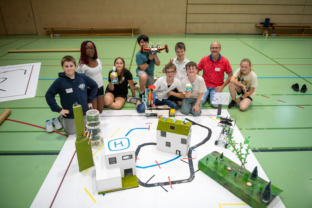 Eine Gruppe von Kindern und zwei Erwachsenen steht auf dem Boden einer Turnhalle. Sie sind um eine Modellstadtanlage mit Spielzeugfahrzeugen, Gebäuden und Bäumen versammelt.