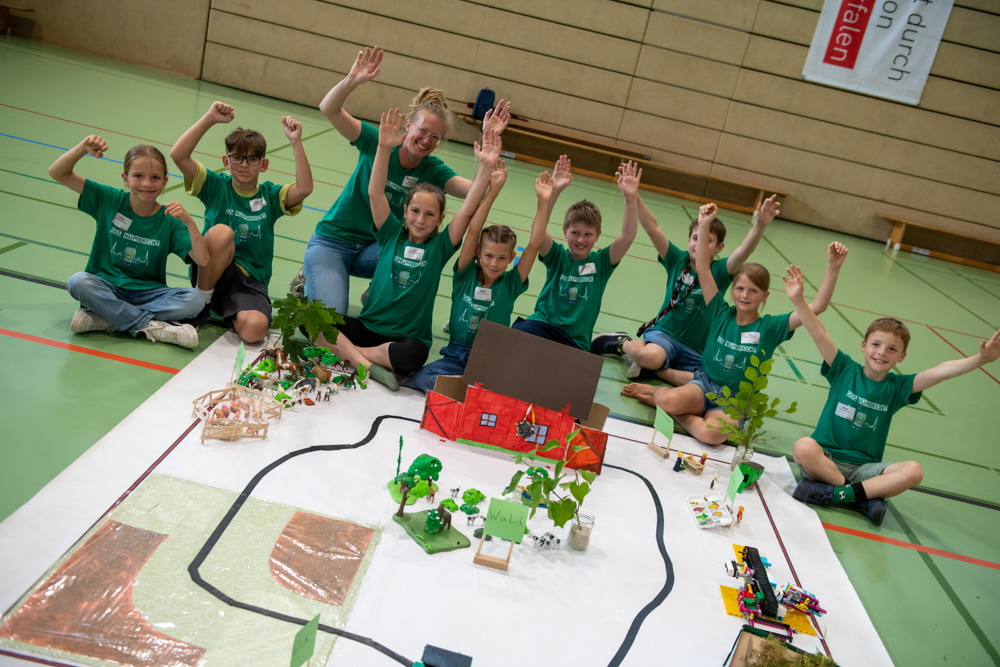 Eine Gruppe von Kindern und ein Erwachsener sitzen um eine große Modellstadt auf dem Boden einer Turnhalle. Sie heben alle ihre Arme und lächeln in die Kamera.