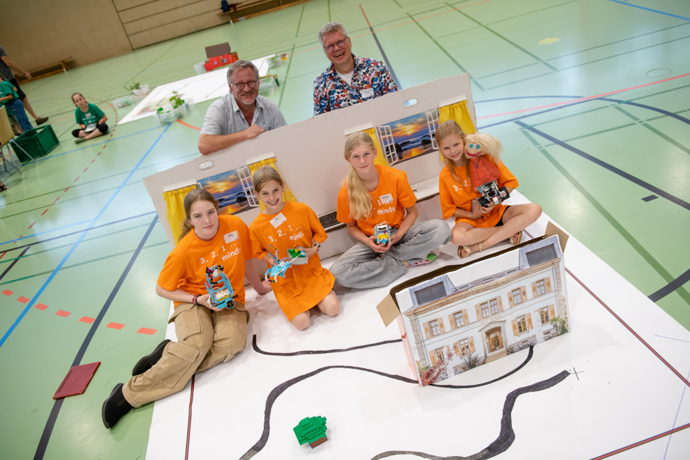 Eine Gruppe von Kindern und Erwachsenen in passenden blauen Hemden posiert in einem Innenraum. Sie stehen hinter einer Auslage mit farbenfrohen Modellgebäuden und Kunsthandwerk, die auf dem Boden angeordnet sind.
