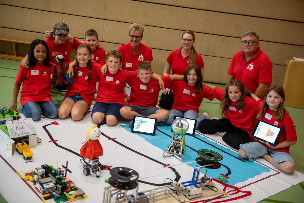 Drei Jungen und ein Erwachsener knien auf dem Boden einer Turnhalle neben einem Modellroboterprojekt. Das Projekt besteht aus beschrifteten Schildern, Spielzeugnahrung, einer grünen Kunstrasenstruktur und Laptops obenauf.