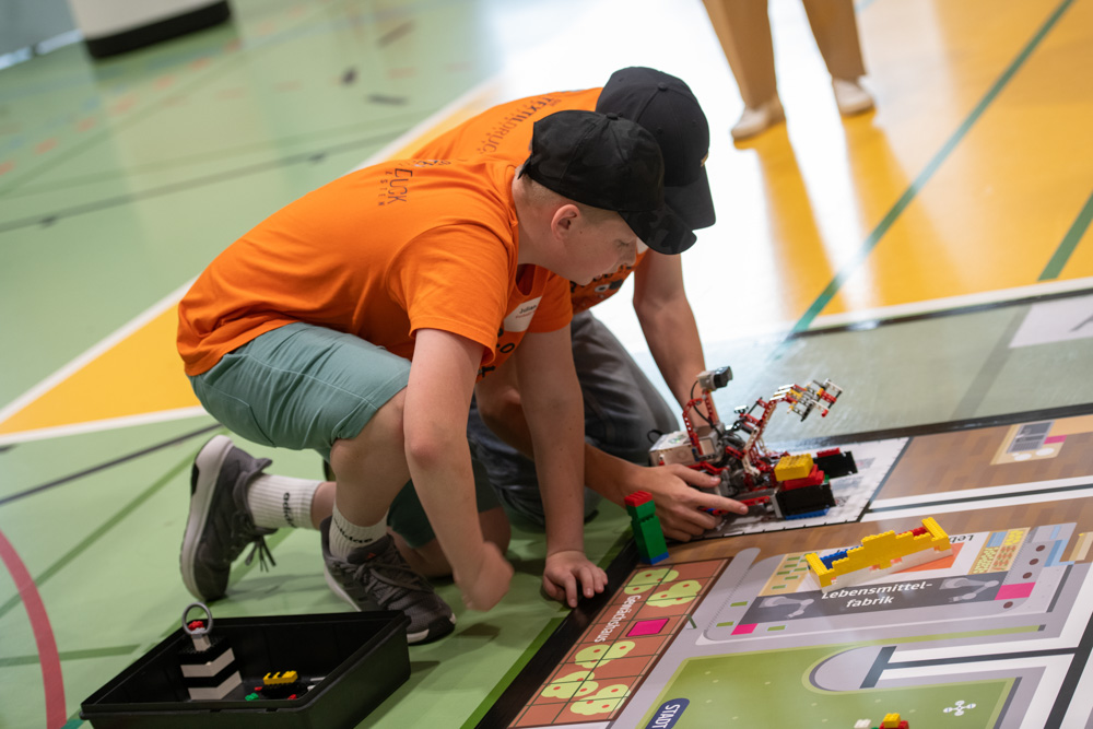 Zwei Jungen knien auf dem Turnhallenboden und arbeiten gemeinsam daran, ein buntes LEGO-Roboterprojekt auf einer Matte während einer Veranstaltung zu bauen oder anzupassen.