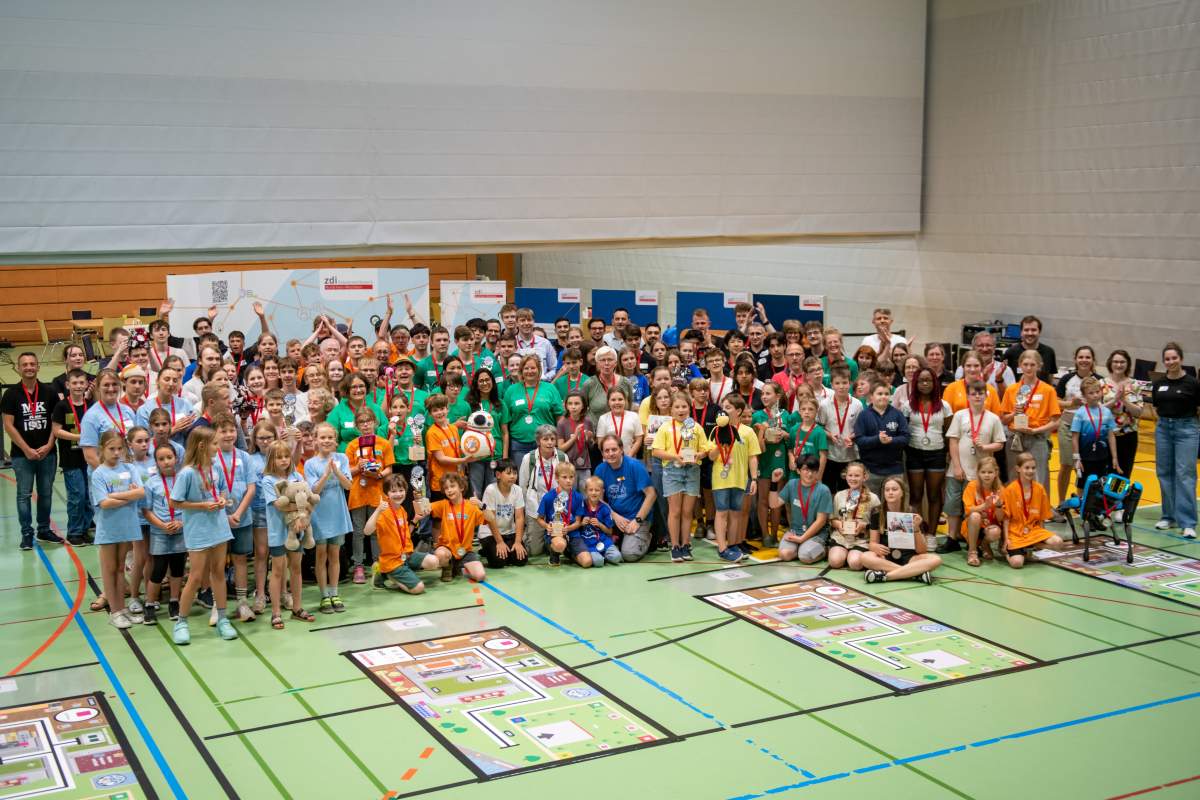 Eine große Gruppe von Erwachsenen und Kindern steht in einer Sporthalle. Im Vordergrund sind die Matten für den zdi-Roboterwettbewerb zu sehen.