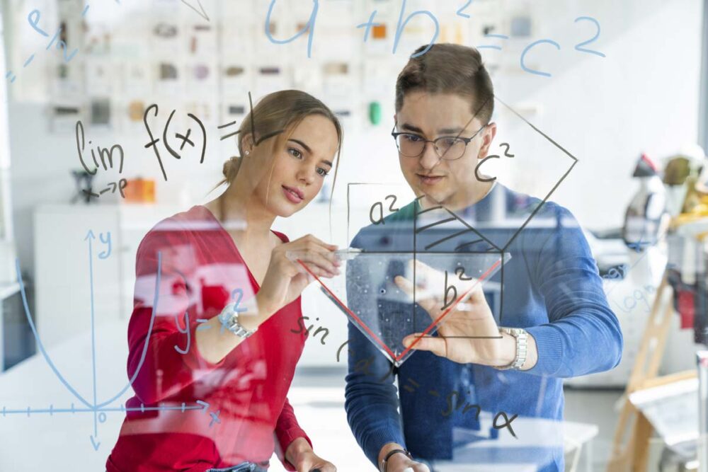 Zwei Personen zeichnen mathematische Graphen und Formeln auf eine transparente Tafel in einem Klassenzimmer oder Labor.