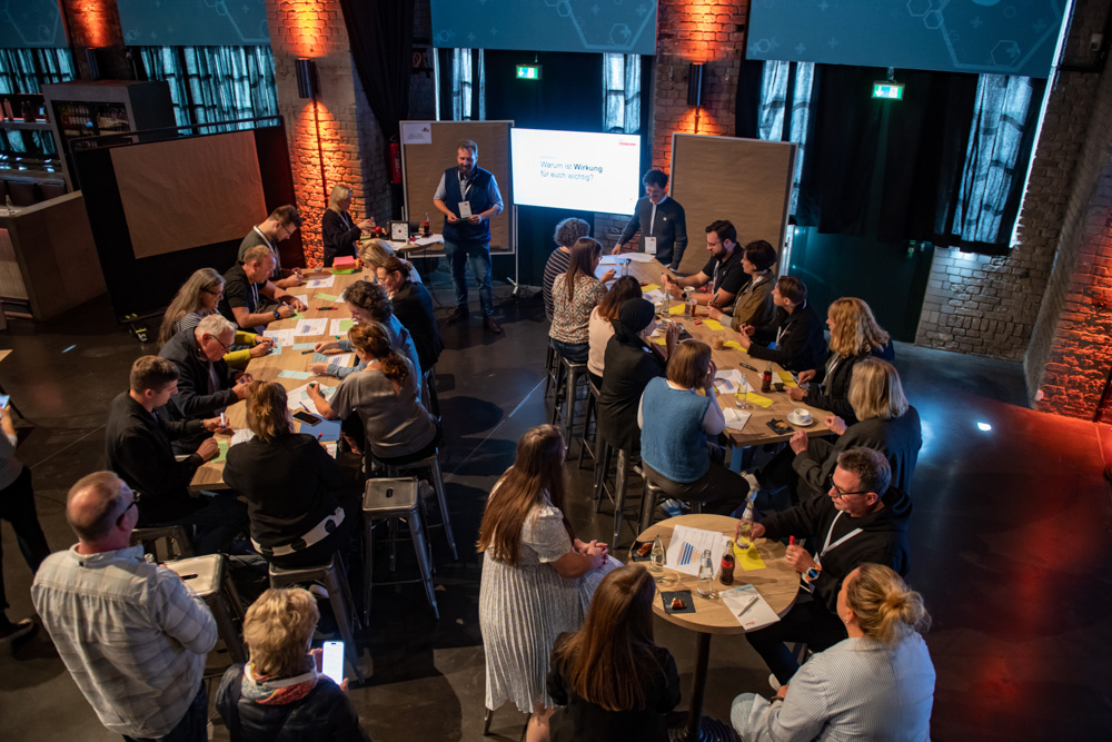 Menschen sitzen an runden Tischen und nehmen an einem Workshop oder einer Diskussion teil. Ein Moderator steht in einem Raum vor einem Bildschirm.