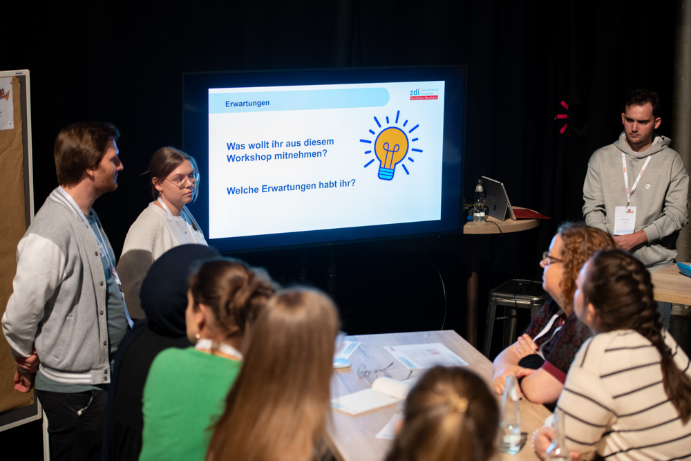 Mehrere Personen sitzen um einen Tisch und diskutieren in einem Workshop. Auf einem großen Bildschirm wird eine Folie mit einer Präsentation über Erwartungen auf Deutsch gezeigt.