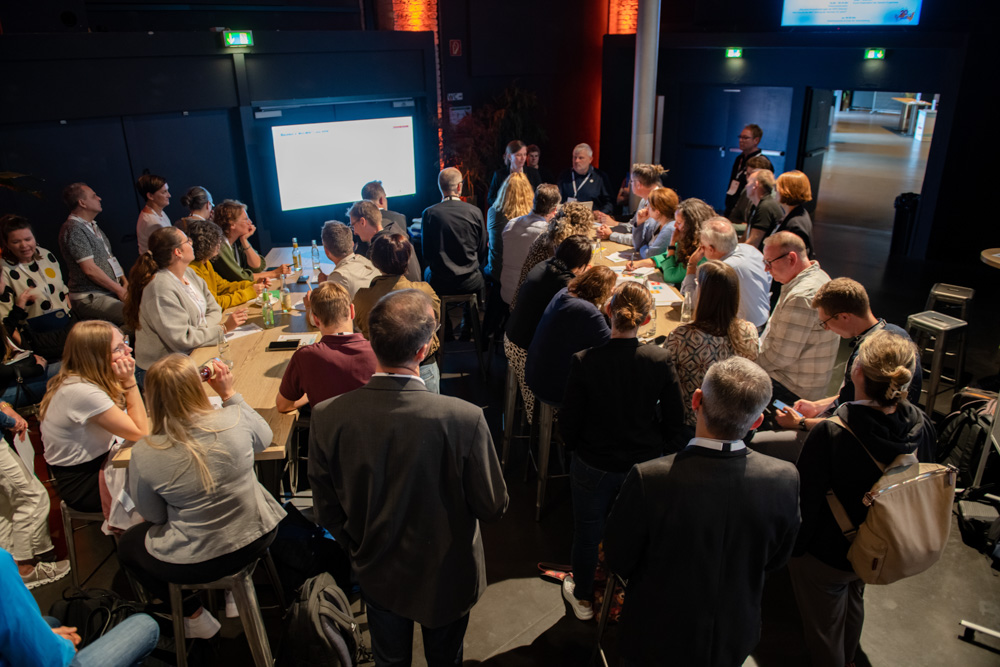 Die Menschen sitzen und stehen in einem Konferenzraum um Tische herum und hören den Rednern zu, die vor einem großen beleuchteten Bildschirm sitzen.