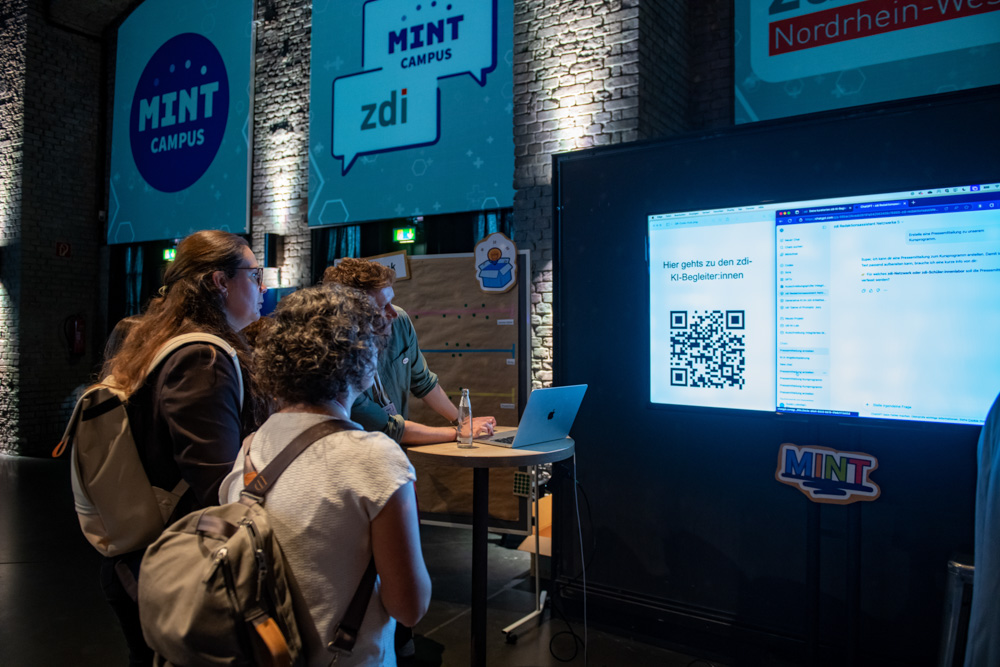 Drei Personen stehen an einem Tisch mit einem Laptop und einem großen Bildschirm, auf dem ein QR-Code angezeigt wird, sowie MINT-Campus-Bannern an der Wand.