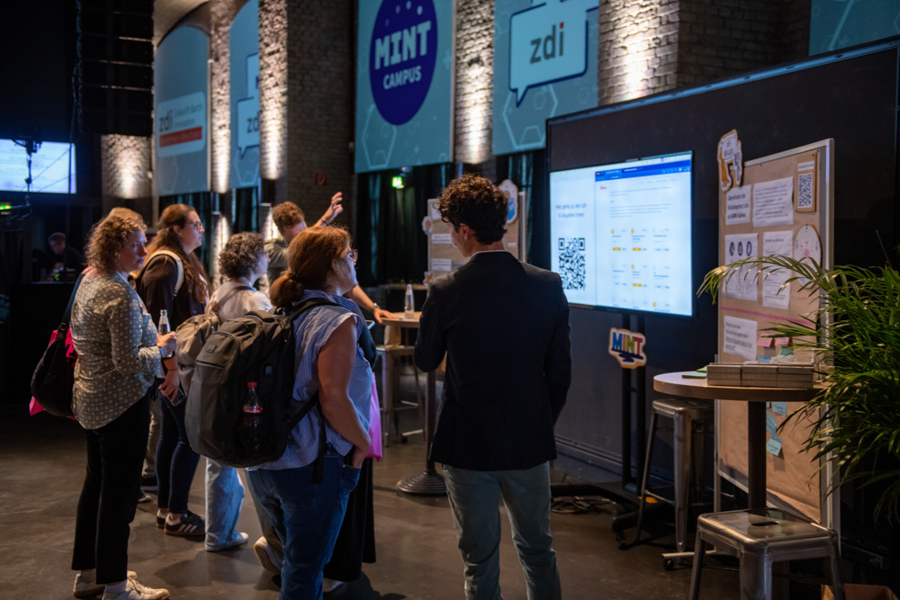 Eine Gruppe von Personen steht bei einer Veranstaltung in einer Halle vor einer digitalen Anzeige und Informationstafeln. An den Wänden hängen Werbebanner.